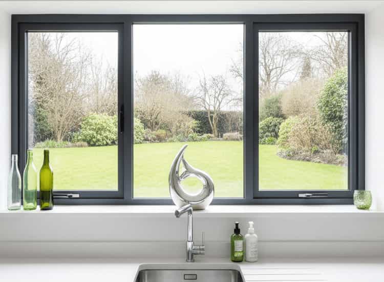 Modern wide grey aluminium window with a sleek frame overlooking a green lawn and garden. The three-panel window is installed above a white kitchen countertop with a stainless steel sink.