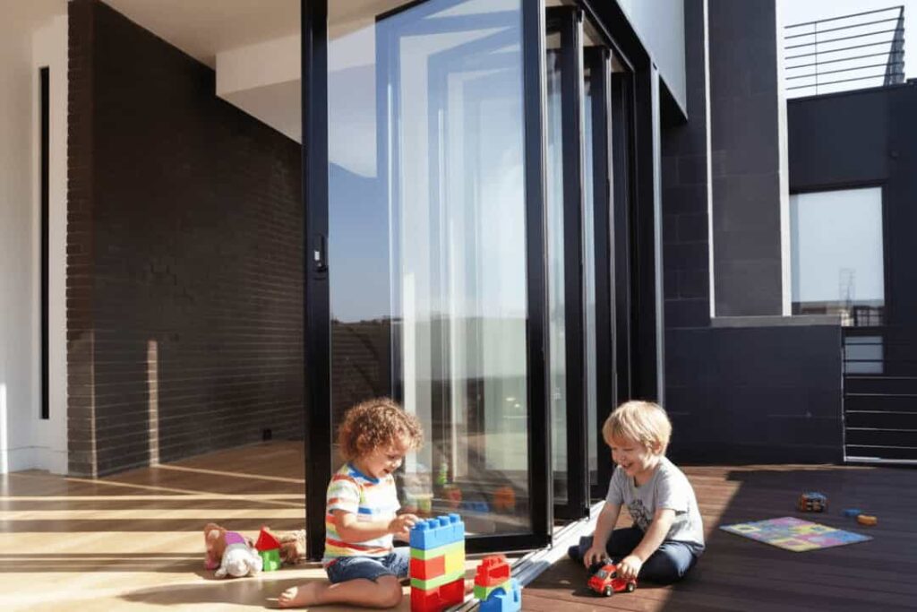 Two young children playing with toys on a wooden deck as an open aluminium bi-fold door connects the outdoor space to the brightly lit indoor living area.