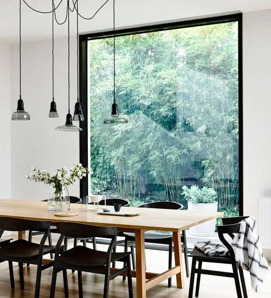 A dining room featuring a large minimalist aluminum fixed picture window overlooking a lush bamboo garden.