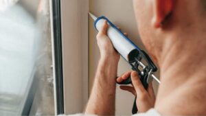 A professional applying a continuous bead of white silicone sealant to the edge of an aluminum window frame to prevent air leaks and water damage.