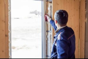 A worker trims spray foam insulation around a white window frame, showing proper sealing to prevent air leaks and drafts.