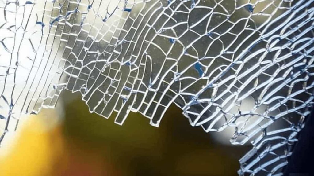 A close-up of a broken tempered glass pane with a central point of impact and radiating spider-web cracks.
