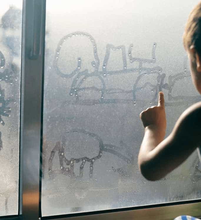 A child's hand drawing a picture with a finger on a foggy glass pane of an aluminium window covered in heavy condensation.