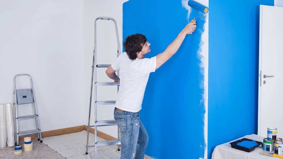 A man in a white t-shirt and jeans stands on a drop cloth, using a paint roller to apply bright blue paint to a room wall next to an aluminum stepladder.