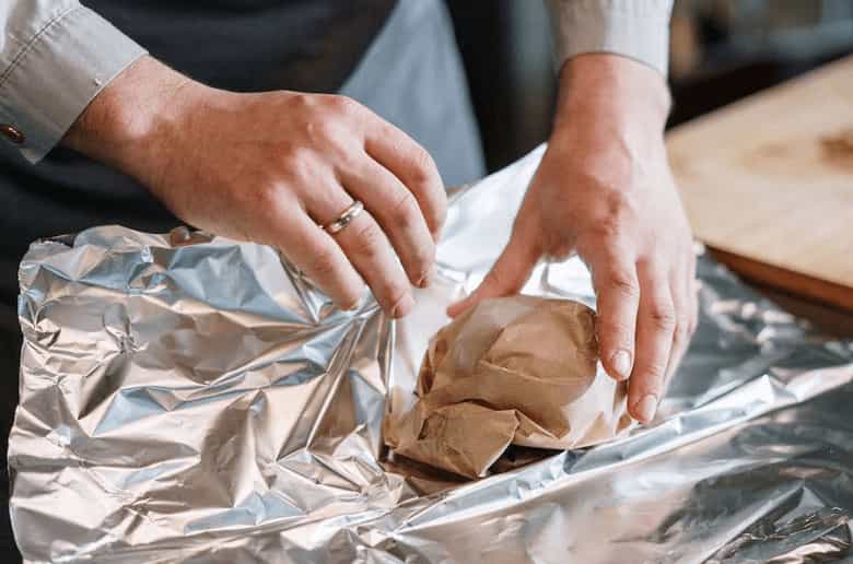 Close-up of hands wrapping a piece of food with a sheet of aluminum foil.