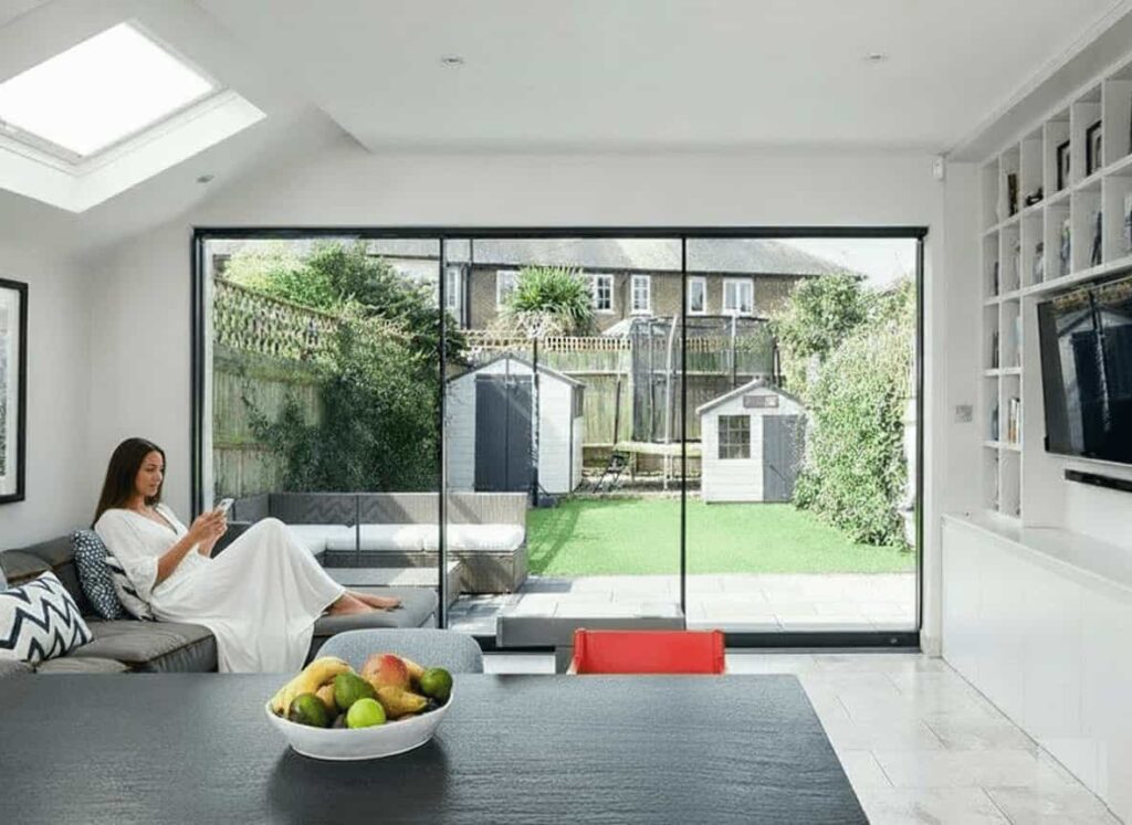 A woman relaxing indoors on a sofa next to a large, slim-framed black aluminium two-track sliding door that overlooks a green backyard and patio.