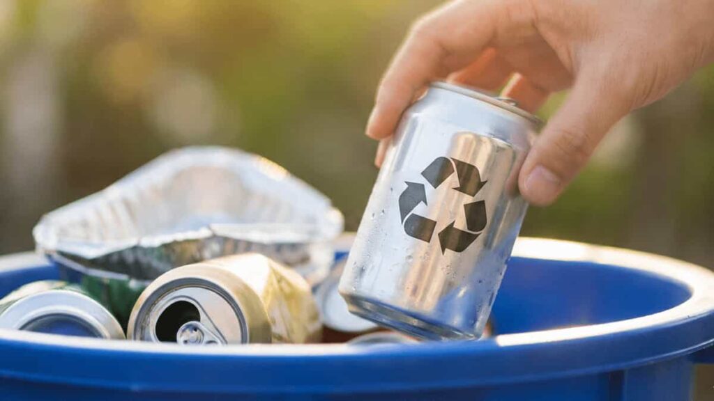 A close-up photograph of a person's hand lowering an empty aluminum beverage can with a visible recycling symbol into a blue plastic recycling bin already containing other cans and foil containers.