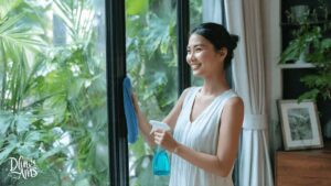 A woman cleaning aluminium window in a bright, naturally lit room with garden view – home maintenance in modern tropical climate.