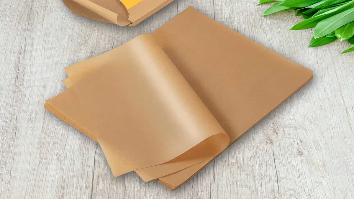Stack of brown parchment paper sheets on a wooden table, next to a box and green leaves, highlighting its use in baking for non-stick cooking.
