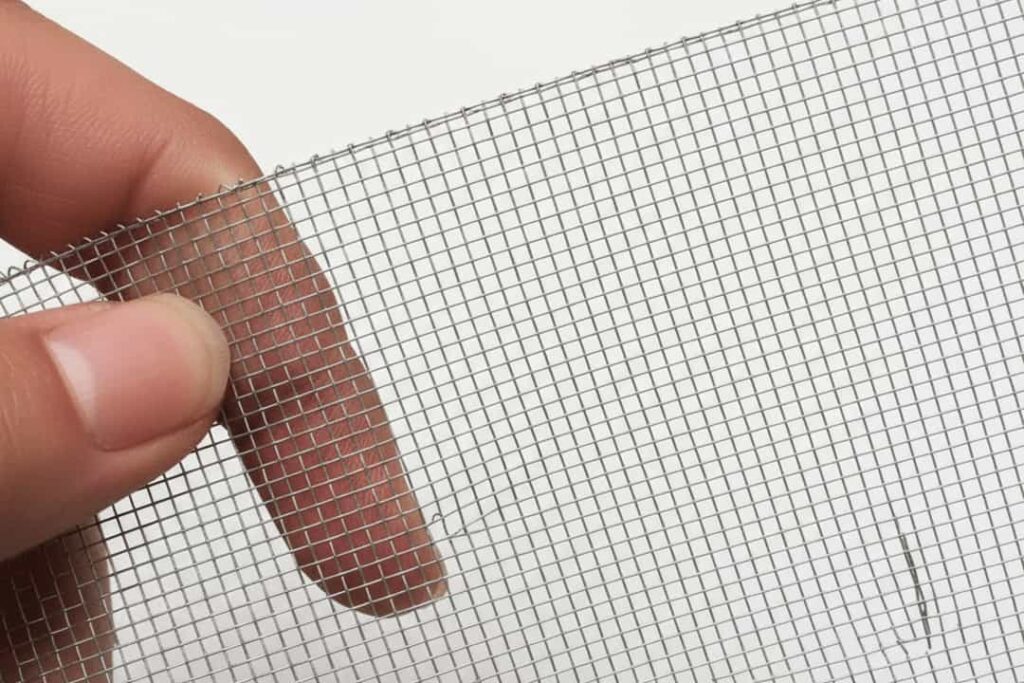 A close-up shot of a hand holding a piece of fine, gray aluminium mesh mosquito net against a white background.