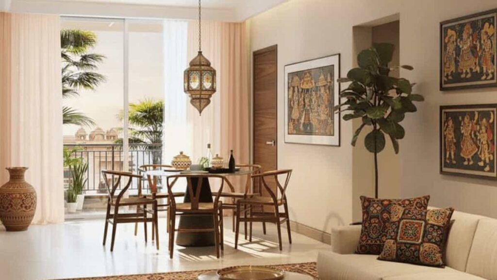 A contemporary living and dining area with strong Indian cultural influences. The space features a cream sofa with patterned cushions, a round wooden dining table with wishbone-style chairs, and multiple framed Indian folk art paintings on the walls, including a row of four on the right. A large window/sliding door opens to a balcony with green plants and a view of a traditional-style building exterior. A decorative Moroccan-style lantern hangs over the dining table, and a terracotta urn stands by the window.