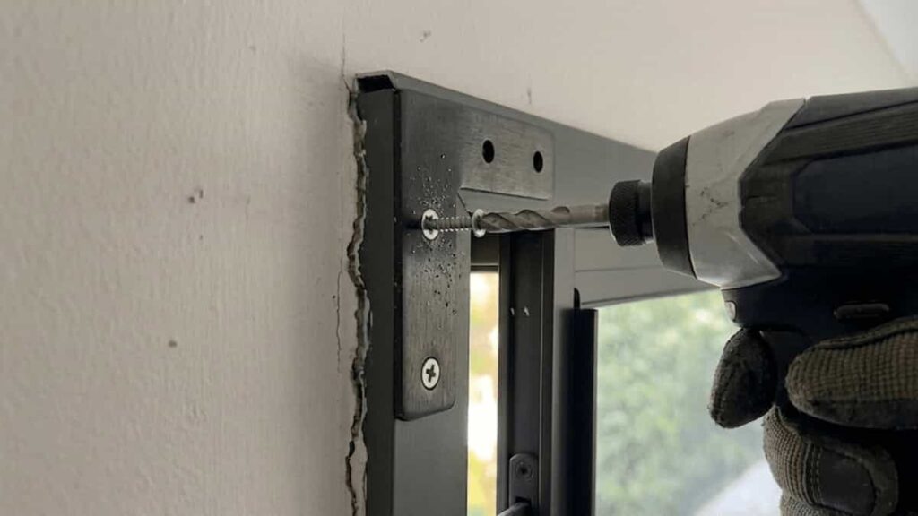 Gloved hand installing a small aluminium corner bracket flush against the inside mitre joint of a dark grey window frame, using a power drill