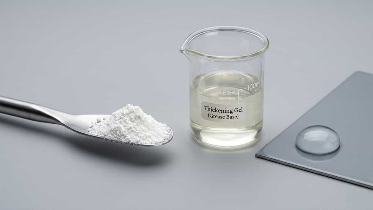 White fluffy aluminum stearate powder on a spatula, with a beaker of thickened oil gel and a water bead on a treated surface.