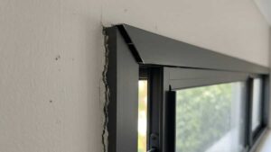 Dark grey aluminium sliding window with a V‑shaped gap at the mitered corner and sash misalignment, indicating frame deformation