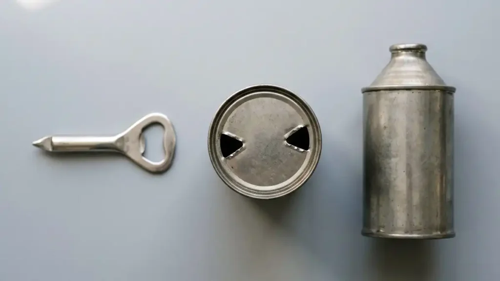 Vintage church key can opener next to a flat-top steel can with two punched holes and a cone-top beer can, showing how cans opened before the pull tab.