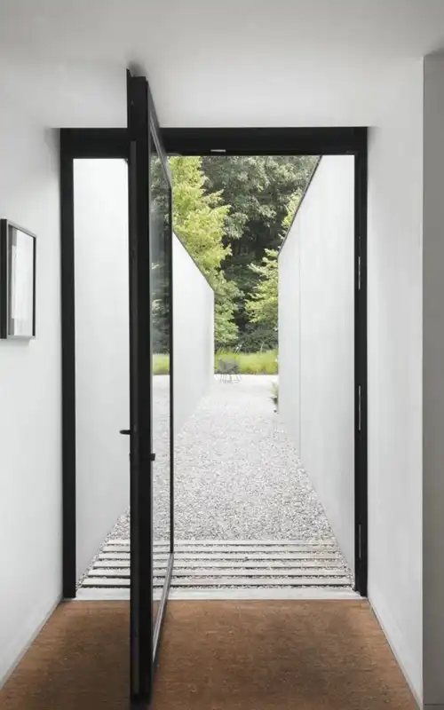 Minimalist aluminum pivot door opening to a gravel patio pathway, perfect for modern indoor-outdoor living.