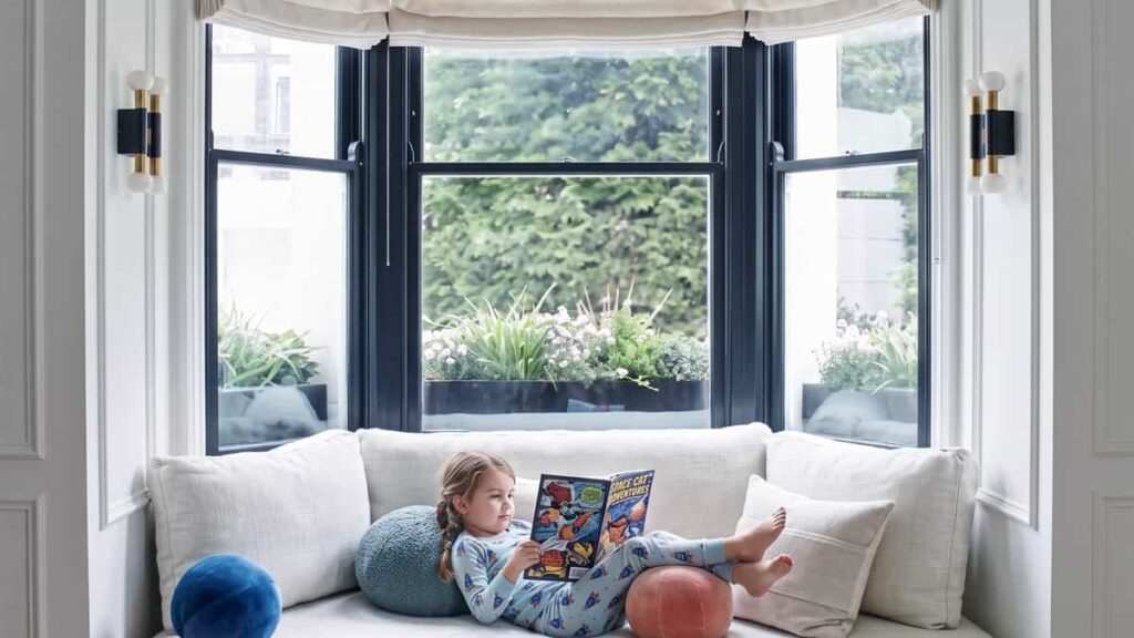 Modern black aluminium bay window with built-in daybed seating and cream Roman shades in a luxury living room